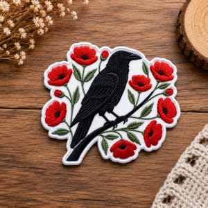 Peut inclure: Un écusson brodé représentant un corbeau noir perché sur une branche sombre, entouré de fleurs de coquelicot rouge vif avec des feuilles et des tiges vertes. Le motif est sur fond blanc avec une bordure blanche festonnée.