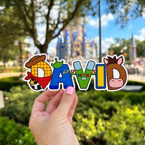 May include: An embroidered patch spelling the name DAVID, where each letter features character-inspired designs including a cowboy hat, a red bandana, a green alien, a space ranger suit, and a brown horse face with a pink snout.