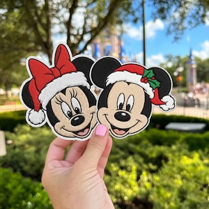 Puede incluir: Dos parches bordados con personajes de ratones de dibujos animados. Un personaje lleva un lazo rojo con borde blanco y el otro un gorro de Papá Noel rojo con borde blanco y hojas de acebo verdes. Ambos tienen orejas negras y cara beige.