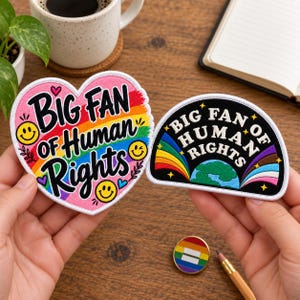 Peut inclure: Deux écussons brodés. Un écusson en forme de cœur présente un fond arc-en-ciel, des visages souriants jaunes et le texte "BIG FAN OF HUMAN RIGHTS" en noir. Le second écusson semi-circulaire a un fond noir, un globe, des rayons arc-en-ciel et le texte "BIG FAN OF HUMAN RIGHTS" en blanc.