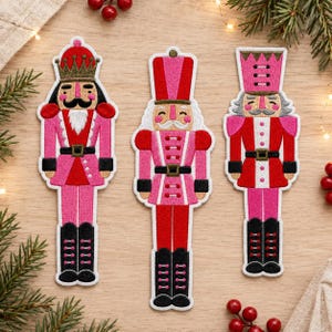 Op de afbeelding: Drie geborduurde notenkraker-patches met roze en rode uniformen en zwarte laarzen. De linkerfiguur draagt een gouden kroon, de middelste figuur een hoge roze hoed en de rechterfiguur een hoge rode hoed met gouden details.