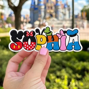 May include: A custom embroidered patch featuring the name SOPHIA in a stylised font. Each letter is designed with distinct character motifs: Mickey Mouse, Minnie Mouse, Pluto, Goofy, a pink bow, and Donald Duck, using red, black, white, yellow, and blue.
