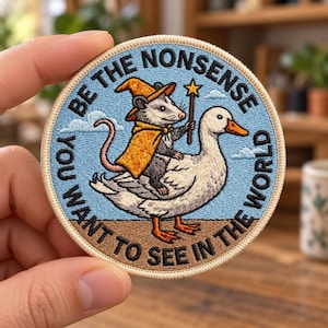 May include: A circular embroidered patch with a light blue background featuring a grey opossum wearing an orange wizard hat and cape, holding a star-tipped wand while riding a white duck. The text reads BE THE NONSENSE YOU WANT TO SEE IN THE WORLD.