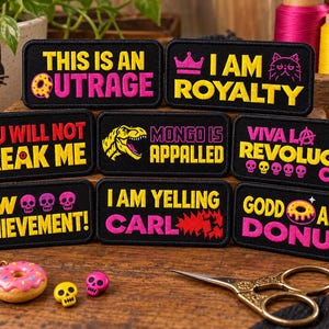 Puede incluir: Una colección de ocho parches rectangulares bordados en negro con texto y gráficos en amarillo y rosa. Las frases incluyen THIS IS AN OUTRAGE, I AM ROYALTY, YOU WILL NOT BREAK ME, MONGO IS APPALLED, VIVA LA REVOLUCIÓN CARL, NEW ACHIEVEMENT, I AM YELLING CARL y GODD AMNIT DONUT.