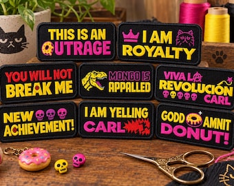 May include: A collection of eight rectangular black embroidered patches with yellow and pink text and graphics. Phrases include THIS IS AN OUTRAGE, I AM ROYALTY, YOU WILL NOT BREAK ME, MONGO IS APPALLED, VIVA LA REVOLUCIÓN CARL, NEW ACHIEVEMENT, I AM YELLING CARL, and GODD AMNIT DONUT.