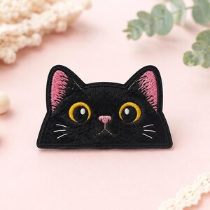 Op de afbeelding: Een geborduurde patch in de vorm van een zwarte kattenkop met grote gele ogen, een kleine roze neus en roze oren. De kat heeft witte geborduurde snorharen op beide wangen en een platte onderkant, uitgevoerd in zwart garen.