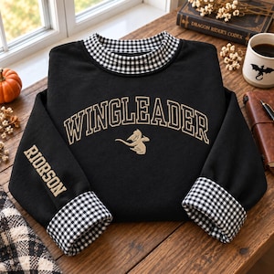 May include: A black long-sleeve sweatshirt featuring a black and white gingham collar and cuffs. The chest displays the text WINGLEADER in gold arched lettering above a small gold dragon graphic. The word RIORSON is embroidered on the left sleeve.