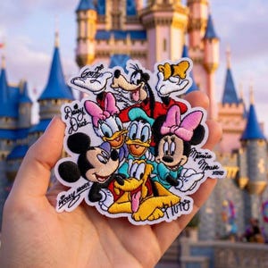 May include: An embroidered patch featuring a group of cartoon characters including Mickey Mouse, Minnie Mouse, Donald Duck, Daisy Duck, Goofy, and Pluto. The patch includes embroidered signatures for each character and the text XOXO.