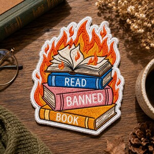May include: An embroidered patch featuring a stack of three books engulfed in orange and yellow flames. The blue book is labelled READ, the pink book is labelled BANNED, and the yellow book is labelled BOOK in white capital letters.