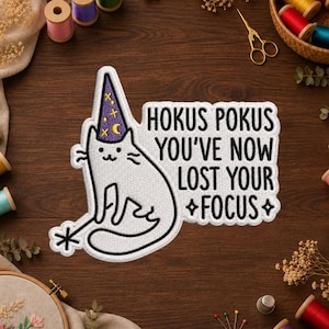 Op de afbeelding: Een witte geborduurde patch met een kat die een paarse tovenaarshoed met gele sterren en manen draagt. De kat houdt een toverstaf vast en de tekst luidt: HOKUS POKUS YOU'VE NOW LOST YOUR FOCUS. Het ontwerp bevat zwarte omlijningen en steraccenten.