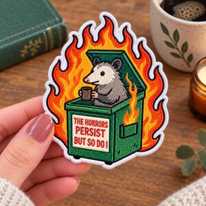 Könnte beinhalten: Ein gestickter Aufnäher mit einem grauen Opossum, das in einem grünen Müllcontainer sitzt, der von hellen orangefarbenen und gelben Flammen umgeben ist. Das Opossum hält eine kleine braune Tasse. Der Text auf dem Container lautet, THE HORRORS PERSIST BUT SO DO I.