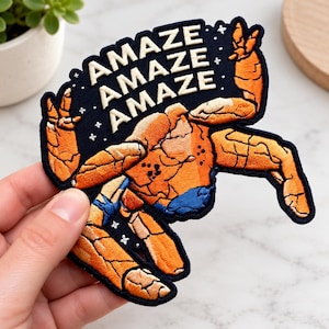 Puede incluir: Un parche bordado que muestra una criatura de piel naranja rocosa con detalles azules sobre un fondo azul marino oscuro. El texto AMAZE AMAZE AMAZE está bordado en hilo crema sobre la figura, rodeado de pequeños detalles en forma de estrella.