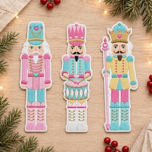 Op de afbeelding: Drie geborduurde notenkraker-patches in pastelkleuren zoals roze, lichtblauw en goud. De linkerfiguur draagt een hoge hoed, de middelste figuur bespeelt een trommel en de rechterfiguur houdt een lange staf vast, allemaal met hartdetails.