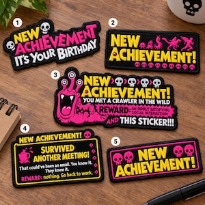 Puede incluir: Cinco parches bordados rectangulares y con formas, con fondo negro y texto en amarillo o rosa. Los diseños incluyen frases de New Achievement, un monstruo rosa, calaveras y textos humorísticos sobre cumpleaños y reuniones.