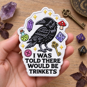 May include: A white embroidered patch featuring a black raven surrounded by a purple crystal, a green potion bottle, a red mushroom, a purple die, a gold key, a pink button, and yellow stars, with the text I WAS TOLD THERE WOULD BE TRINKETS.