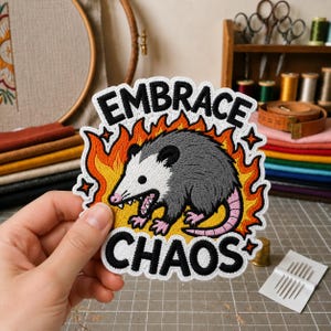 May include: An embroidered patch featuring a gray and white opossum with a pink nose and tail, surrounded by orange and yellow flames. The text EMBRACE CHAOS is stitched in bold black letters above and below the opossum on a white background.