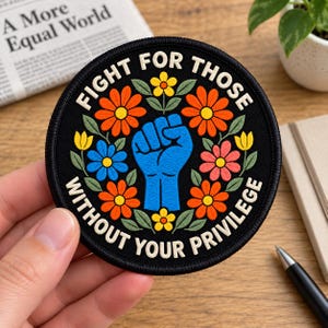 May include: A circular embroidered patch with a black background featuring a central blue raised fist surrounded by colorful orange, yellow, pink, and blue flowers with green leaves. The text reads FIGHT FOR THOSE WITHOUT YOUR PRIVILEGE.