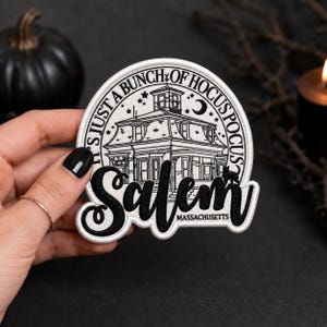 Peut inclure: Un écusson brodé circulaire présentant une illustration en noir et blanc d'une maison avec le texte IT'S JUST A BUNCH OF HOCUS POCUS, Salem et MASSACHUSETTS. Le motif inclut un croissant de lune, des étoiles et une silhouette de chat noir.