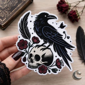 Peut inclure: Un écusson brodé représentant un corbeau noir perché sur un crâne humain beige. Le motif comprend des fleurs rouge foncé, une plume noire unique et de petits points noirs sur un fond blanc avec un bord brodé blanc.
