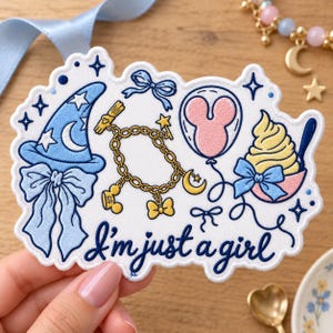 May include: A white embroidered patch featuring blue, yellow, and pink thread designs including a wizard hat, a charm bracelet, a balloon, and a soft serve treat. The text "I'm just a girl" is embroidered in dark blue cursive script at the bottom.
