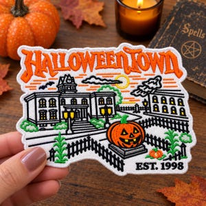 May include: An embroidered patch featuring the text HALLOWEENTOWN in orange, a white building with black outlines, a jack-o'-lantern on a pedestal, a black fence, green plants, and the text EST. 1998 in black thread on a white fabric background.