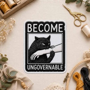 May include: A rectangular embroidered patch with a black border featuring a black cat biting a human arm. The text BECOME is printed in white above the cat and UNGOVERNABLE is printed in white below the cat, all on a white background.