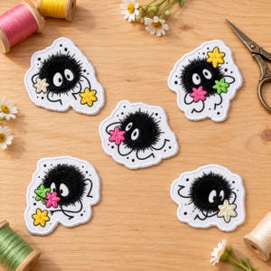 Può includere: Cinque toppe ricamate raffiguranti creature nere, rotonde e pelose con grandi occhi bianchi. Ogni toppa ha un bordo bianco ed è decorata con piccoli fiori colorati nelle tonalità del giallo, rosa e verde, disposti su una superficie.