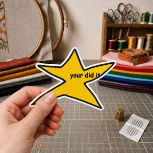 Puede incluir: Un parche bordado en forma de estrella amarilla con un borde negro y el texto negro "your did it" impreso en el centro. El parche presenta una superficie de tela texturizada y se sostiene frente a un fondo de diversos materiales de costura.