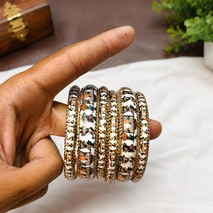 May include: A set of five bangles featuring a white base with intricate, multi-coloured hand-painted figures of people in traditional attire. Each bangle is bordered by thin, textured gold-tone metallic edges, creating a cohesive and detailed design.