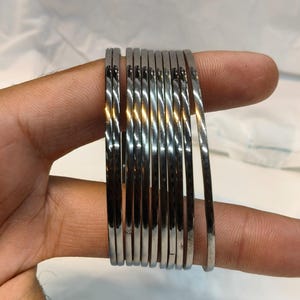 Può includere: Un set di nove bracciali rigidi sottili in metallo color argento lucido con una superficie liscia e riflettente. I bracciali circolari sono impilati insieme, mostrando un design uniforme ed essenziale con una finitura brillante.