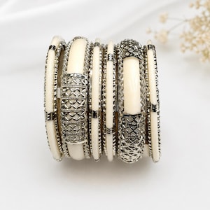 Op de afbeelding: Een stapel van meerdere armbanden met afwisselend gladde, crèmekleurige banden en ingewikkelde zilverkleurige metalen segmenten met een kruislings rasterpatroon. De set bevat dunne effen banden en bredere decoratieve stukken met texturen.
