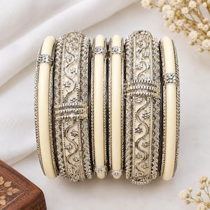 Può includere: Un set di sei bracciali rigidi con design alternati di fasce lisce color crema e fasce in metallo argentato con intricati motivi floreali a volute e bordi testurizzati. I bracciali sono impilati, mostrando una combinazione di motivi semplici e decorati.