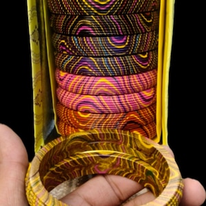 May include: A collection of circular bangles featuring intricate, multi-coloured wavy line patterns. The set includes stacked bangles in shades of red, yellow, green, purple, pink, and orange, presented in a yellow box with two loose bangles.