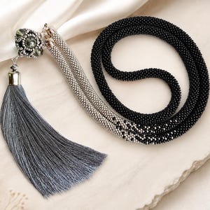 Puede incluir: Un collar largo con una cadena de cuentas negras y plateadas que transiciona a una sección negra sólida. El collar incluye una cuenta central con incrustaciones de cristal, una tapa plateada y una borla larga de seda de color gris.