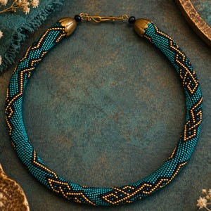 May include: A circular beaded necklace featuring a repeating geometric pattern in teal and black glass beads. The necklace is finished with gold-toned metal end caps and a gold-toned hook clasp, creating a continuous, structured, and tubular design.