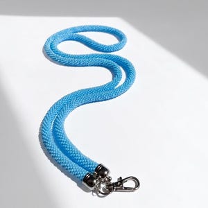 Puede incluir: Un cordón de cuentas de color azul cielo brillante, elaborado con pequeñas cuentas de vidrio uniformes tejidas en un tubo flexible similar a una cuerda. Los extremos están asegurados con tapas de metal plateado y un mosquetón.