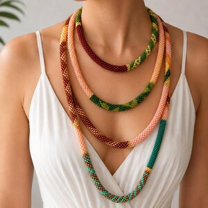 Puede incluir: Un collar largo de varias hebras de cuentas con una paleta de colores vibrantes que incluye rojo oscuro, verde bosque, melocotón suave, bronce y turquesa. El collar está hecho de cuentas diminutas y uniformes tejidas en una estructura tubular.
