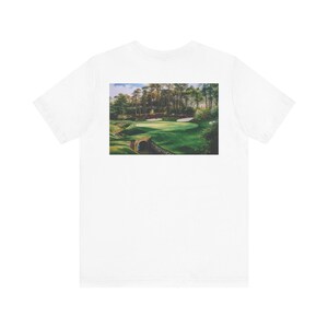 May include: White t-shirt with a landscape painting of a golf course. The artwork shows a green golf course with trees, sand traps, and a bridge. The shirt is made of a soft, comfortable material.