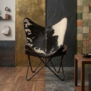 May include: A black and white cowhide butterfly chair with a black metal frame. The chair is set against a backdrop of a gold and black wall, with a wooden floor. The chair is a stylish piece of furniture.
