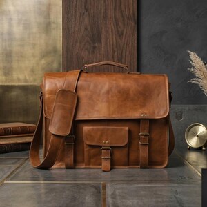 May include: A brown leather messenger bag with a shoulder strap, handle, and front pocket. The bag has buckles and is set against a backdrop of wood and a dark wall. A small gold clock and books are also visible.
