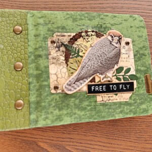 Puede incluir: Un diario verde con lomo texturizado y tres tachuelas de latón. La cubierta muestra un collage con una ilustración de halcón, una hoja verde recortada y una etiqueta negra con el texto blanco "FREE TO FLY" sujeto por un clip de metal.