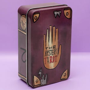 May include: A rectangular purple metal tin featuring a graphic of a tan hand with six fingers. The text "THE MYSTERY TAROT" is printed in black and red on the palm. The tin has illustrated corner accents and a circular stamp on the side.