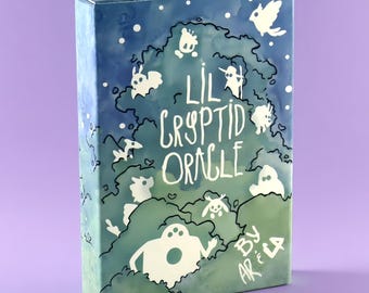Puede incluir: Una caja de cartas rectangular para el Lil Cryptid Oracle de Ariela, con un degradado estilo acuarela de azul a verde. El frente muestra siluetas blancas de varias criaturas y el texto Lil Cryptid Oracle by Ariela.