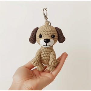 May include: A small, tan, crocheted dog keychain with dark brown ears, black bead eyes, and a white muzzle featuring a black plastic nose and stitched mouth. The figure is attached to a silver-toned metal lobster clasp at the top of its head.