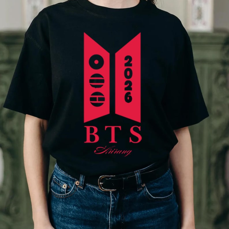 May include: A black short-sleeved t-shirt featuring a red graphic design on the chest. The design includes two vertical trapezoids, the text 2026, the letters BTS, and the word Arirang in a script font, all printed in a bold red colour.
