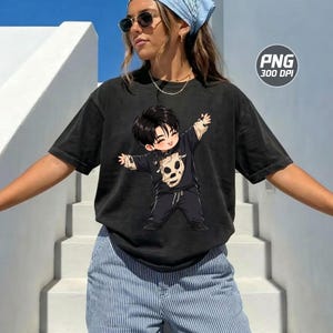 May include: A black short-sleeve t-shirt featuring a graphic of a cartoon character with dark hair wearing a black shirt with a beige skull print. The character has arms outstretched and is wearing black trousers. The shirt has a crew neckline.