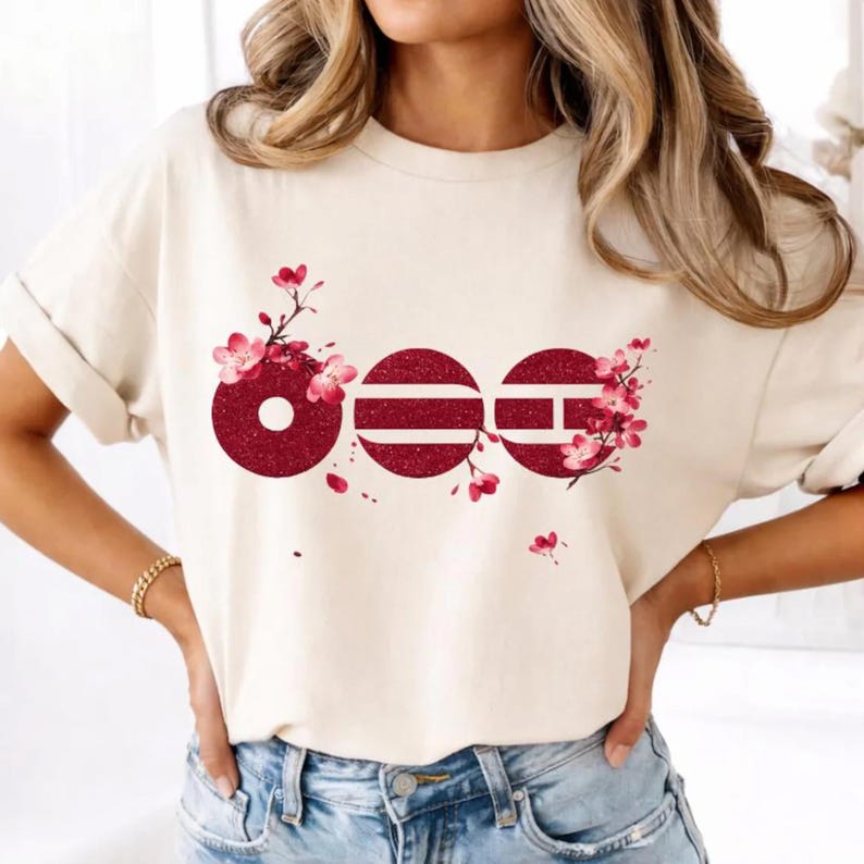 May include: A cream-coloured short-sleeve t-shirt featuring a graphic design of three circular shapes in a dark red glitter finish. The circles are adorned with delicate pink cherry blossom branches and flowers, creating a floral pattern.