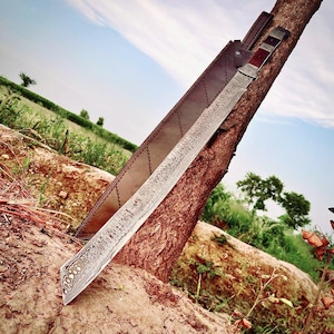 May include: A large, ornate sword with a patterned blade and a brown leather sheath. The sword is resting against a tree trunk, with a rural landscape in the background. The handle features red and silver accents.