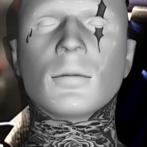 May include: A white mannequin head with closed eyes and black tear and star-shaped markings. The neck features a detailed black and white tattoo of a rose, skulls, and other floral elements. The overall aesthetic is gothic.
