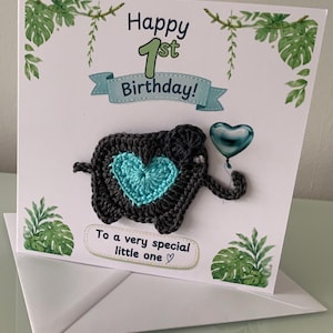 May include: A square greeting card featuring a dark grey crocheted elephant with a bright turquoise heart on its side, holding a metallic blue heart-shaped balloon. The card displays the text Happy 1st Birthday and To a very special little one.